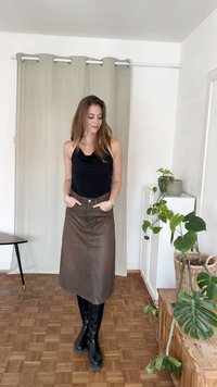 Saia midi de couro marrom com um leve brilho, botões frontais e bolsos laterais. Combinada com uma blusa preta sem mangas e botas pretas até ao joelho.