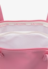 Růžová otevřená taška s bílou síťovanou vnitřní kapsou a vnitřním lemováním s nápisem "LACOSTE" v růžovém textu.
