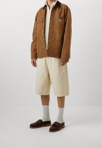 Giacca di tela marrone con colletto a camicia, tasche frontali e bottoni, abbinata a pantaloncini crema, calzini grigi e scarpe marrone scuro.