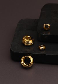 Ensemble de bijoux en or composé de boucles d'oreilles créoles texturées et d'une bague avec des accents ondulés, présentés sur des surfaces en pierres foncées.