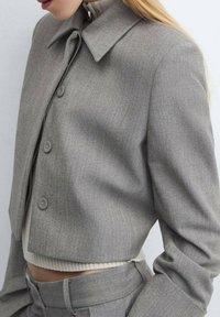 Chaqueta gris recortada con cuello puntiagudo, tejido texturizado y cierre frontal con botones, combinada con un suéter acanalado de color claro debajo.