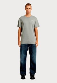 Lichtgrijs katoenen T-shirt met korte mouwen, met een klein logo op de linkerborst. Gecombineerd met donkerblauwe wijde spijkerbroek en zwarte schoenen.