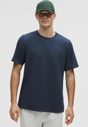 Mand iført en mørkeblå kortærmet t-shirt, lysegrå bukser og en mørkegrøn baseballkasket, som vender fremad mod en ensfarvet baggrund.