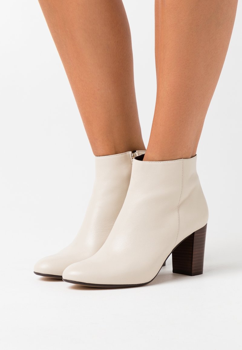 Bottes blanches en cuir montantes à la cheville avec talons blocs, portées sur des jambes nues sur un fond blanc uni.