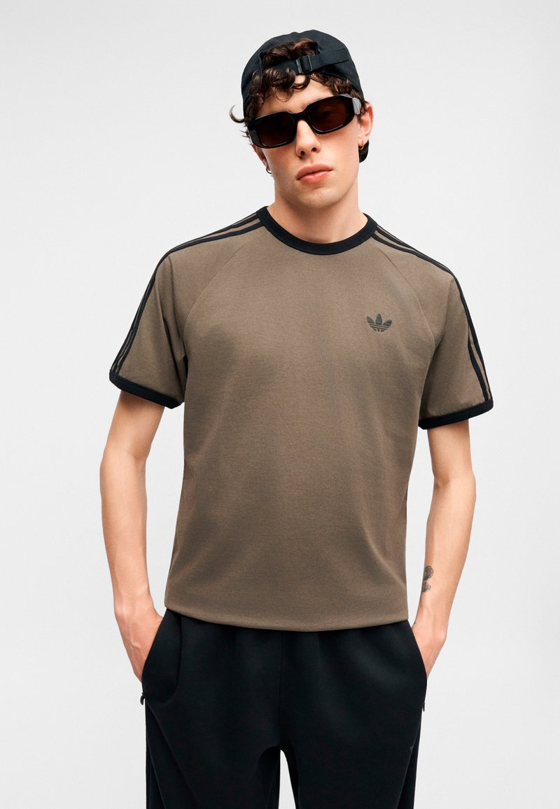 Ung mand iført solbriller, sort kasket, olivengrøn Adidas T-shirt med sorte striber og sorte bukser, stående med hænderne i lommerne.