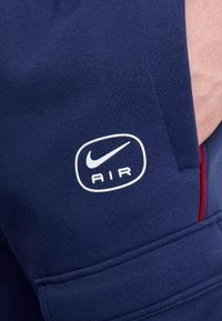 Närbild av marinblå Nike Air-byxor med vit logotyp och röd kant vid en hand i fickan.