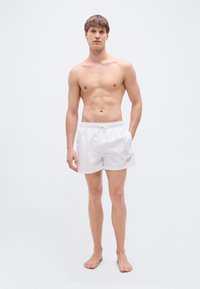 Άσπρα μαγιό σορτς από ελαφρύ υλικό, με κορδόνι στη μέση, μήκος πάνω από το γόνατο και διακριτικό λογότυπο στο πλάι.