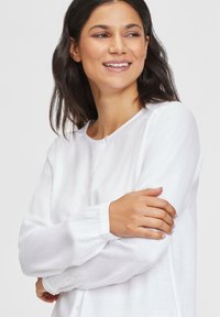 Weiße Langarmbluse mit rundem Ausschnitt und Knopfdetails. Aus leichtem Stoff gefertigt, mit gerafften Manschetten und glatter Textur.