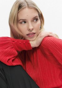 Rød plisseret top med tekstureret finish og knapdetalje på manchet, kombineret med en sort underdel. Neutral baggrund.