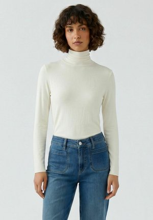 Vrouw met krullend haar die een wit coltrui met lange mouwen en een hoge taille blauwe spijkerbroek draagt, staat tegen een effen lichte achtergrond.