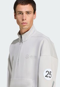 Lys grå zip-up pullover med høj krave, frontlomme, broderet logo og en rund applikation med nummeret 25 på ærmet.