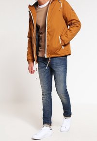 Orange hættejakke med hvid lynlås og snore. Lys brun sweater under, parret med blå jeans og hvide sneakers.