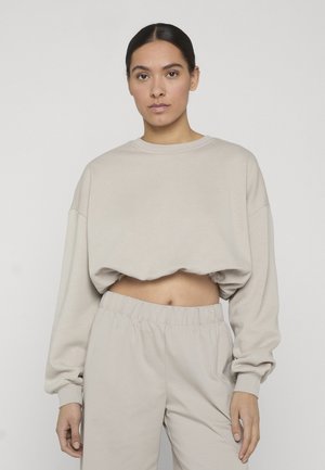 Femme aux cheveux foncés attachés, portant un sweat-shirt court beige et un pantalon assorti à taille élastique, sur un fond uni.