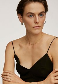 Vestido negro sin mangas con escote en V, con finas tirantes. Modelo con accesorios de collar dorado y pendientes de aro. Textura suave.