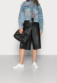 Kvinna som bär en grafisk jeansjacka, grå polotröja, svarta lädershorts till knäna, vita sneakers och håller en svart quiltad kuvertväska.