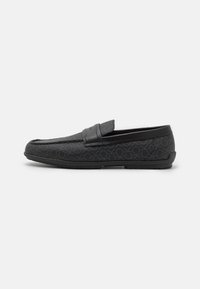 Mocassins noirs avec un tissu texturé arborant un motif subtil, une bride en cuir lisse sur le dessus, et une semelle en caoutchouc flexible.