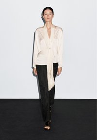 Blusa di seta beige chiaro con un nodo drappeggiato, indossata con pantaloni in suede neri e sandali neri con cinturini, che mettono in risalto texture morbide e tonalità neutre.