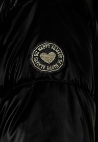 Geacă puffer neagră cu un patch brodat rotund care prezintă un design cu inimă și textul "HAPPY KIDS" aurit pe margine. Textură moale.