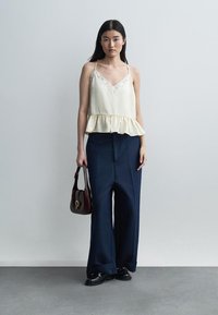 Nainen, joka pukeutunut kermaan vivahtavaan pitsireunuksiseen hihattomaan puseroon, laajalahkeisiin tummansinisiin housuihin ja mustiin kenkiin, pitäen tummanruskeaa käsilaukkua, seisoo yksiväristä harmaata taustaa vasten.