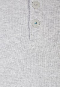 Tkanina jasnoszarej koszulki polo o teksturowanym wzorze dzianiny, z trzema guzikami przy kołnierzu i widocznym przeszyciem.