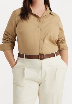 Kobieta w beżowej koszuli na guziki z podwiniętymi rękawami, włożona w białe spodnie z wysokim stanem i brązowym paskiem, ręce w kieszeniach.