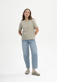 Hellgrünes Baumwoll-T-Shirt, lockere Passform, runder Halsausschnitt. Kombiniert mit hellblauen Jeans in gerader Passform und grauen Schlupfschuhen.