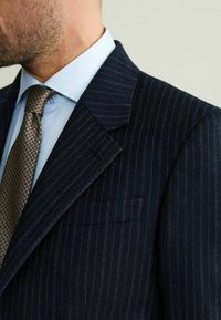 Granatowa marynarka w prążki z klapami o stałym kształcie, teksturowanym materiałem i kieszenią na piersi, zestawiona z jasnoniebieską koszulą i brązowym krawatem w wzory.