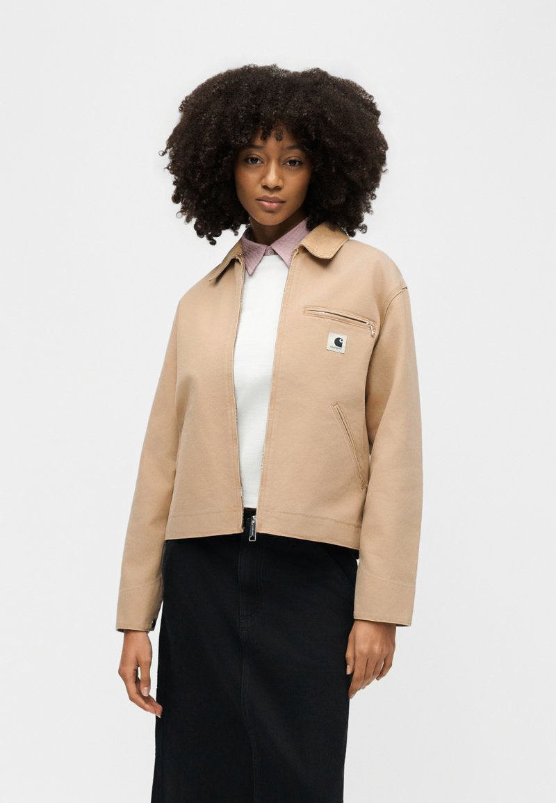 Woman with curly hair wearing a tan zip-front jacket over a white shirt and black skirt, standing against a plain light background.