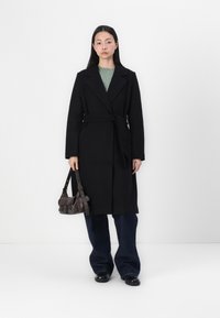 Manteau en laine noir avec une ceinture, revers en pointu et manches longues, associé à un pull en tricot vert, un jean en denim foncé évasé et un sac marron.