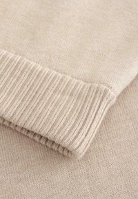 Beige gestrickter Pulloverärmel mit geripptem Bunddetail, der eine weiche Textur und feine Stiche zeigt.