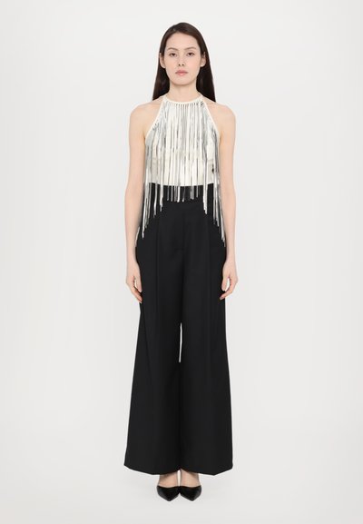 Top halter argintiu cu detalii lungi de franjuri, asortat cu pantaloni negri cu talie înaltă și croială largă. Modelul stă pe un fundal neutru.