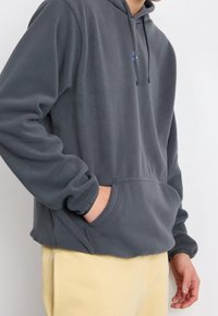 Grå fleece-hoodie med en känguruficka och justerbar huva med dragsko. Har en liten blå logga på bröstet.