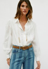 Hvid skjorte med knaplukning og broderede detaljer, puffede ærmer, båret med højtaljede blå stribede jeans og et brunt bælte. Guld lagdelt halskæde.