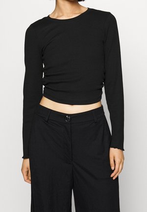 Personne portant un top court noir moulant à manches longues et un pantalon noir taille haute avec fermeture à bouton.