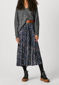 Grauer gestrickter Pullover mit V-Ausschnitt, kombiniert mit einem blauen Maxirock mit floralen Mustern und gestuftem Design. Dunkle Stiefeletten mit dicken Sohlen.