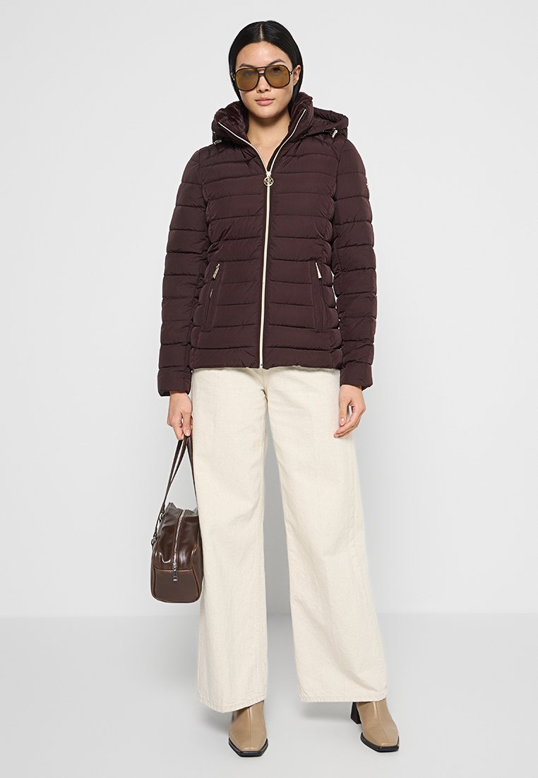 Doudoune marron foncé avec capuche, fermeture éclair blanche contrastante et deux poches latérales, associée à un pantalon large de couleur claire et des lunettes de soleil.