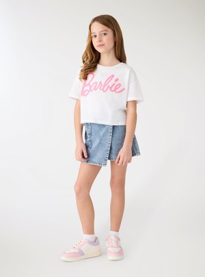 Ragazza giovane in piedi che indossa una t-shirt bianca "Barbie", gonna di jeans e sneakers bianche con dettagli rosa e viola su uno sfondo bianco semplice.