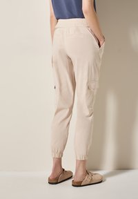 Pantalons cargo en coton beige avec taille et poignets élastiques ; poches latérales et poches à rabat sur les jambes. Portés avec des sabots marron clair.