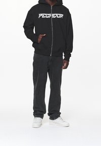 Sudadera negra con cremallera y "PEGADOR" impreso en blanco, combinada con jeans oscuros y zapatillas blancas. La tela parece suave con un ajuste casual.