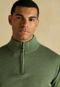 Jeune homme aux cheveux courts et bouclés, portant un pull vert à demi-fermeture éclair avec un col côtelé, debout devant un fond beige uni.