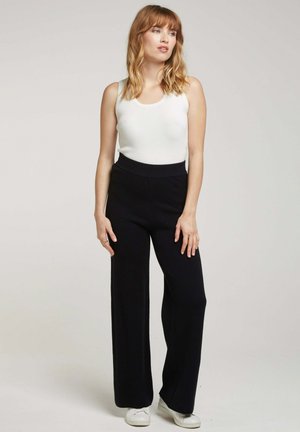 Pantalons larges noirs en tissu doux, associés à un débardeur blanc ajusté. Le modèle porte des baskets blanches et se tient les mains sur les hanches.