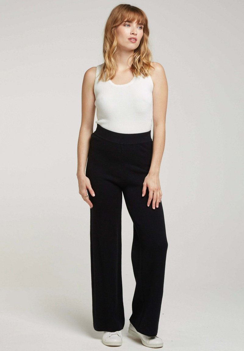 Pantalons larges noirs en tissu doux, associés à un débardeur blanc ajusté. Le modèle porte des baskets blanches et se tient les mains sur les hanches.