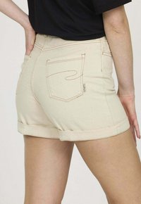 Shorts en denim beige clair avec ourlets retroussés, comportant deux poches arrière avec des coutures contrastées et une petite étiquette logo près de la poche droite.
