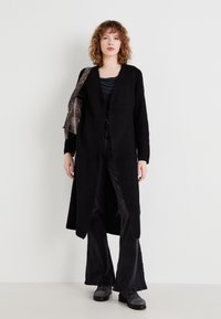 Cardigan lung negru cu un tricot texturat, cu deschidere frontală, purtat cu un top negru și blugi evazați, completat cu cizme negre până la gleznă și o geantă maro.