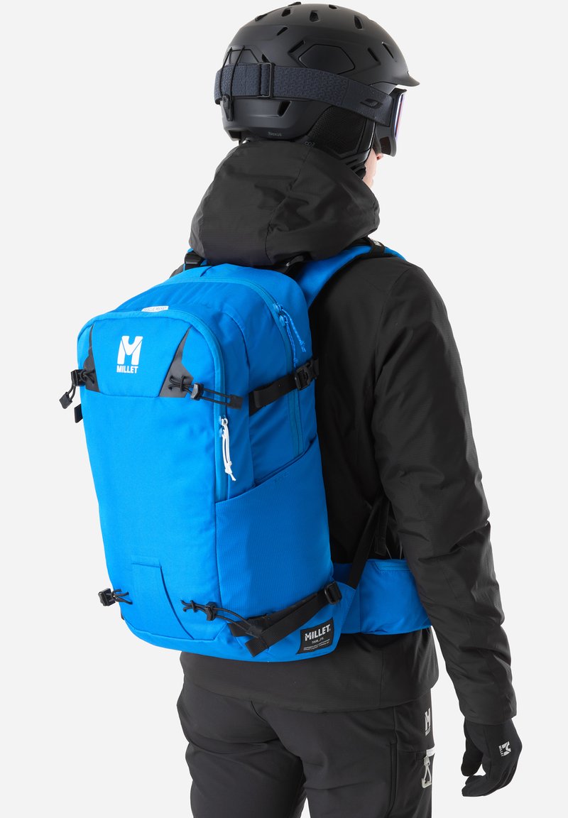 Personne portant une tenue de ski noire, un casque, des lunettes de protection et un sac à dos bleu vif Millet avec des bretelles rembourrées et plusieurs compartiments.