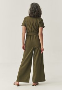 Olijfgroene jumpsuit met korte mouwen, wijde pijpen en een riem om de taille. Gemaakt van zachte, gestructureerde stof. Eenvoudig ontwerp met een sluiting aan de achterkant.