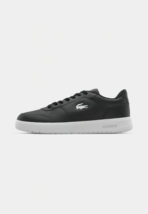 Baskets en cuir noir avec semelle en caoutchouc blanche, présentant un bout rond, un design à lacets et le logo Lacoste sur le côté. Texture lisse.