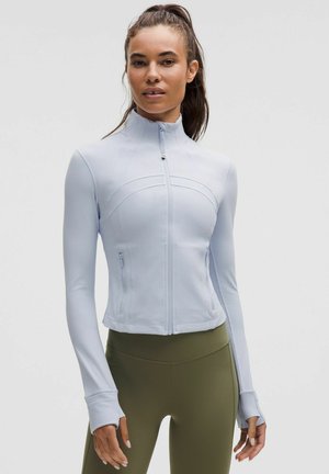 Woman with dark hair in a ponytail wears a light blue zip-up jacket with thumb holes and olive green high-waisted leggings.