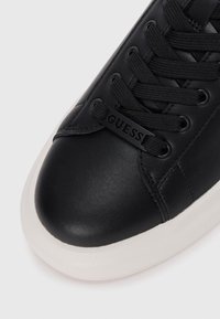 Black leather sneaker with black laces, rounded toe cap, and white rubber sole. "GUESS" logo is embossed on the side.
