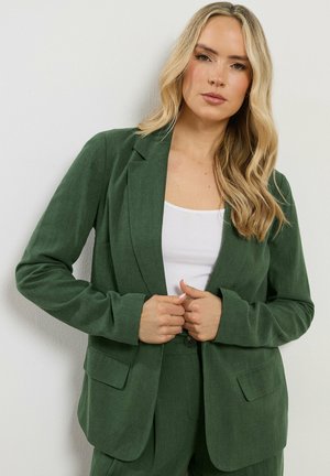 Blonde vrouw die een donkergroene blazer en broek draagt over een witte top, staand tegen een effen witte achtergrond.